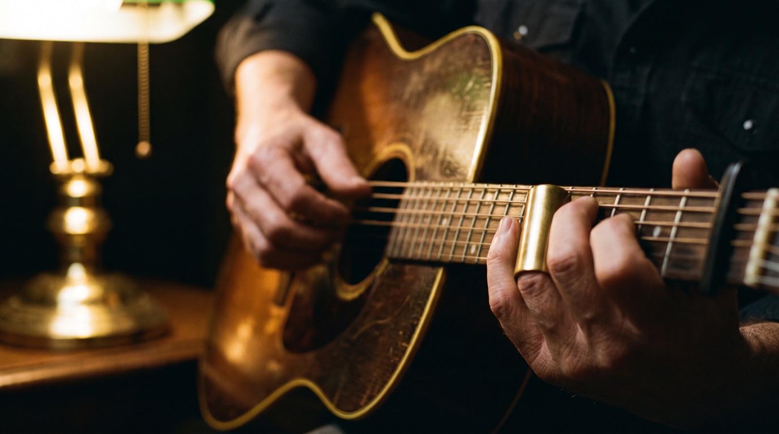 Close-up of hands playing an acoustic guitar near a brass lamp